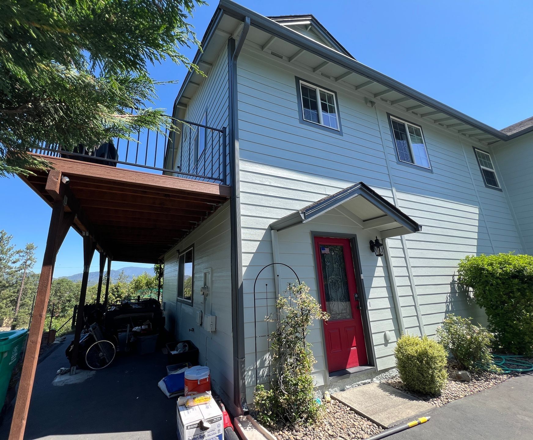 A two-story light blue house with a red front door, a covered porch, and a second-story balcony on a sunny day.