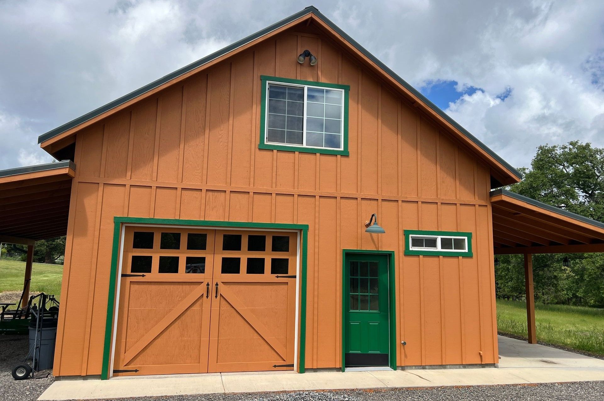 A rust-colored barn with green trim, a large wooden garage door, a single green door, and two windows set in a grassy field.