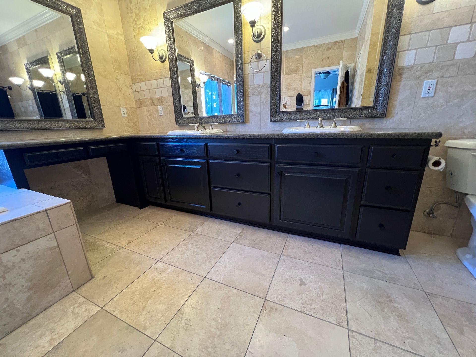 A spacious bathroom with a long, dark wood vanity, dual sinks, ornate mirrors, and beige stone tile flooring and walls.