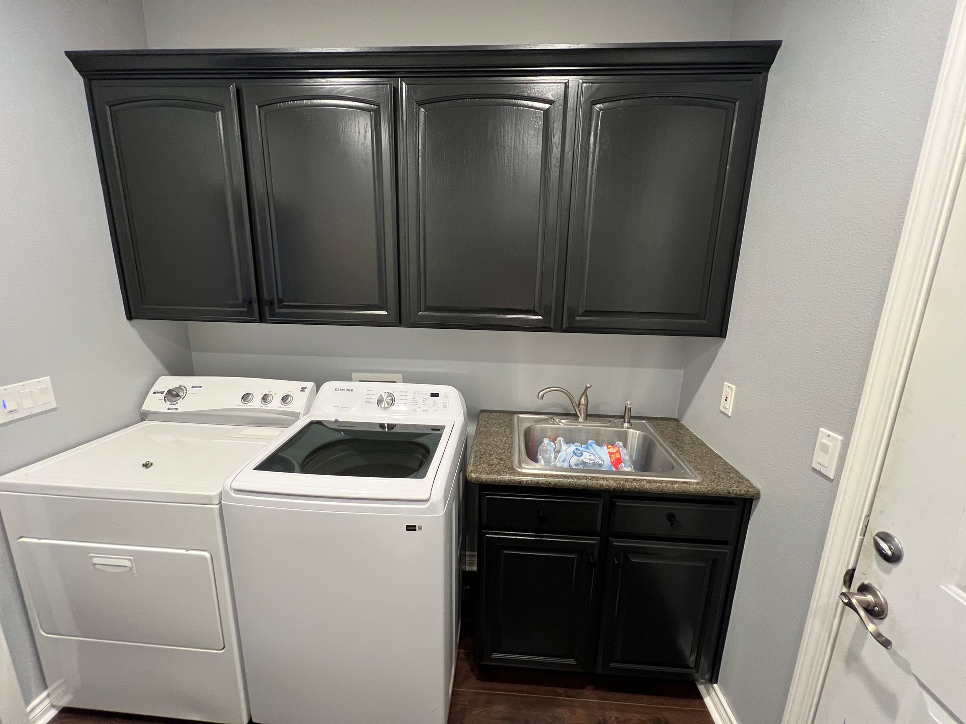 A laundry room with a white dryer and washer next to a small sink with dark cabinets above and below.