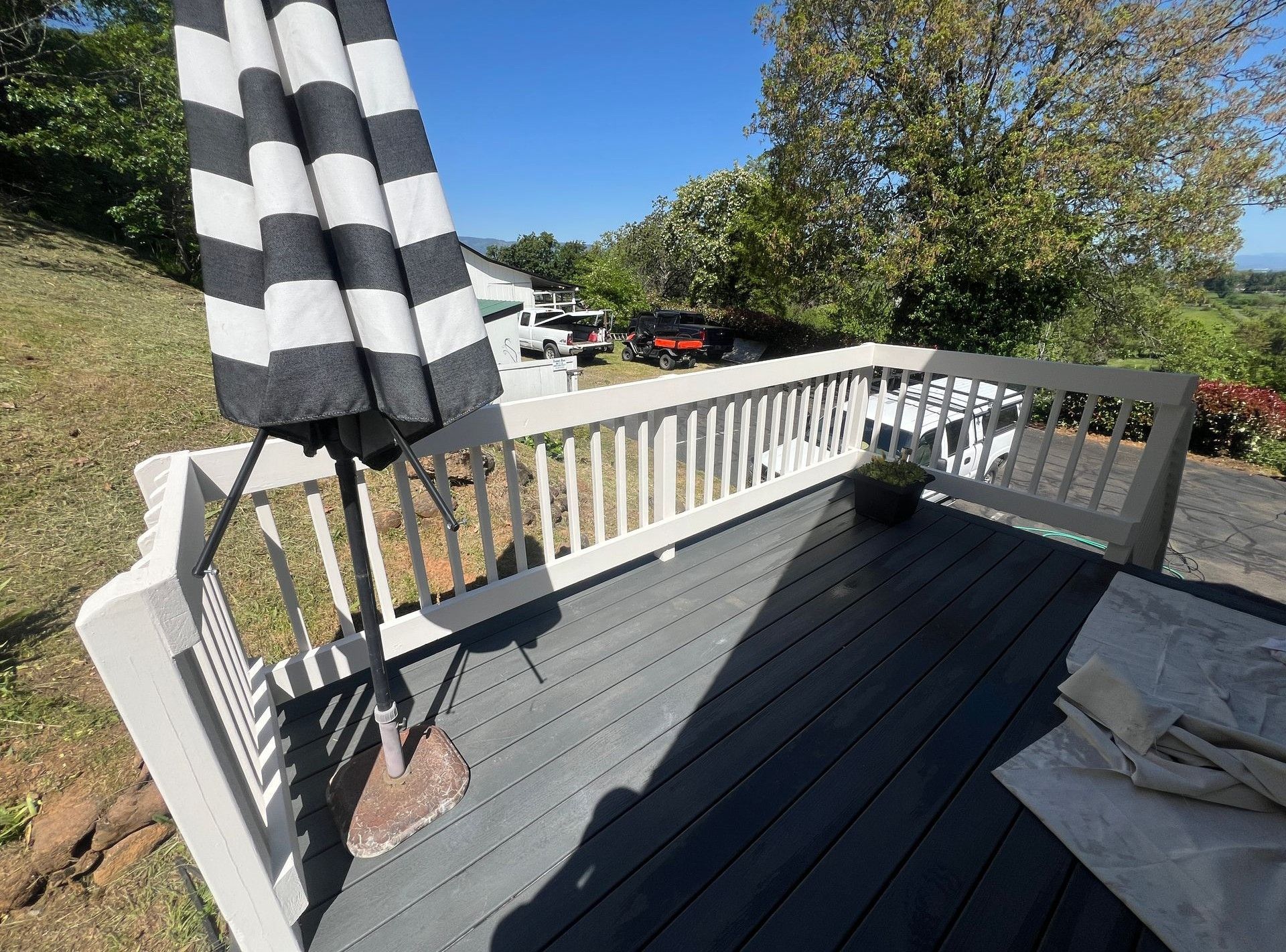 A raised wooden deck with a black and white striped umbrella, white railings, and a view of trees and parked vehicles.