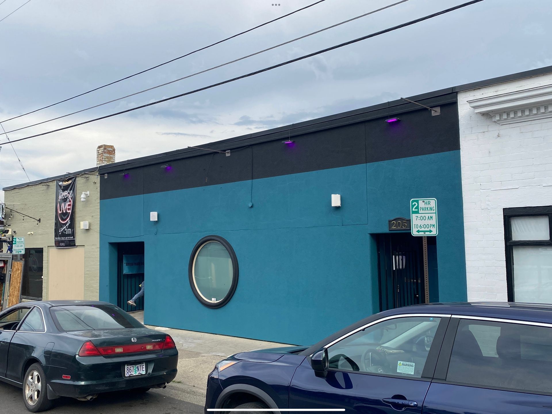 A teal, flat-roofed building on a city street with a circular central window, flanked by two cars parked at the curb.