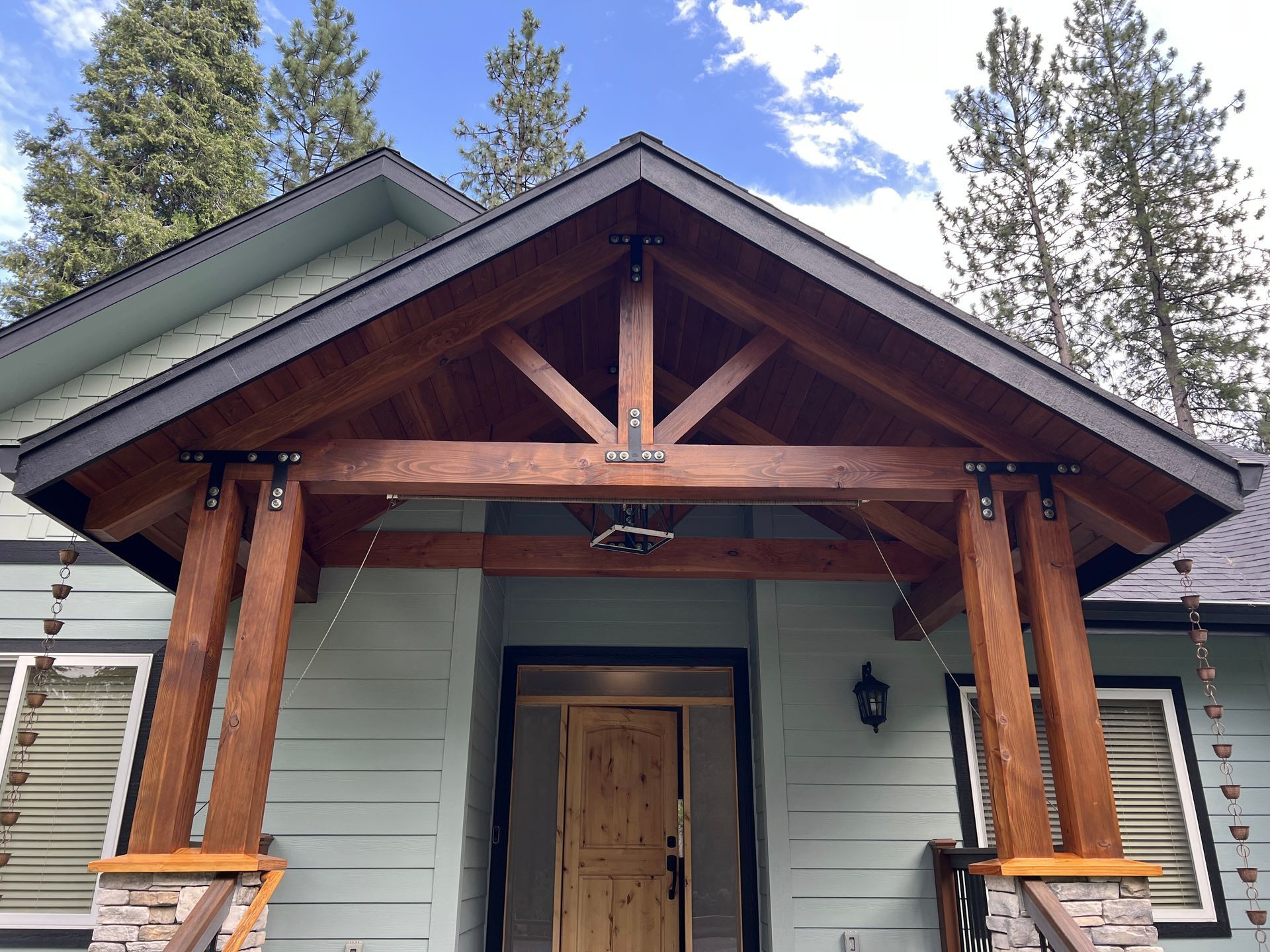 A light green house with a rustic, stained-wood timber-framed porch entryway and stone pedestal columns.
