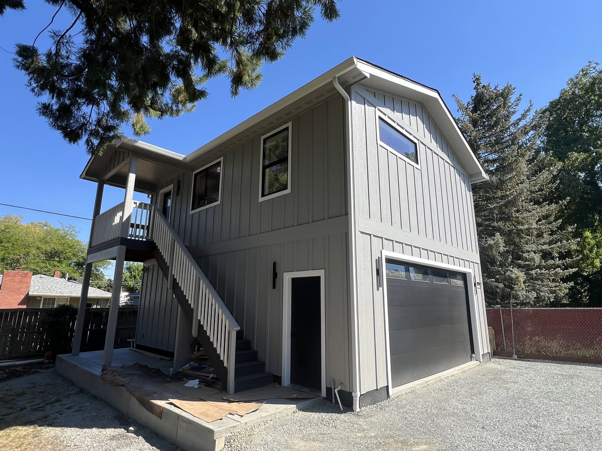 A grey two-story garage with a black door, a gravel driveway, and an exterior staircase leading to an upper level.