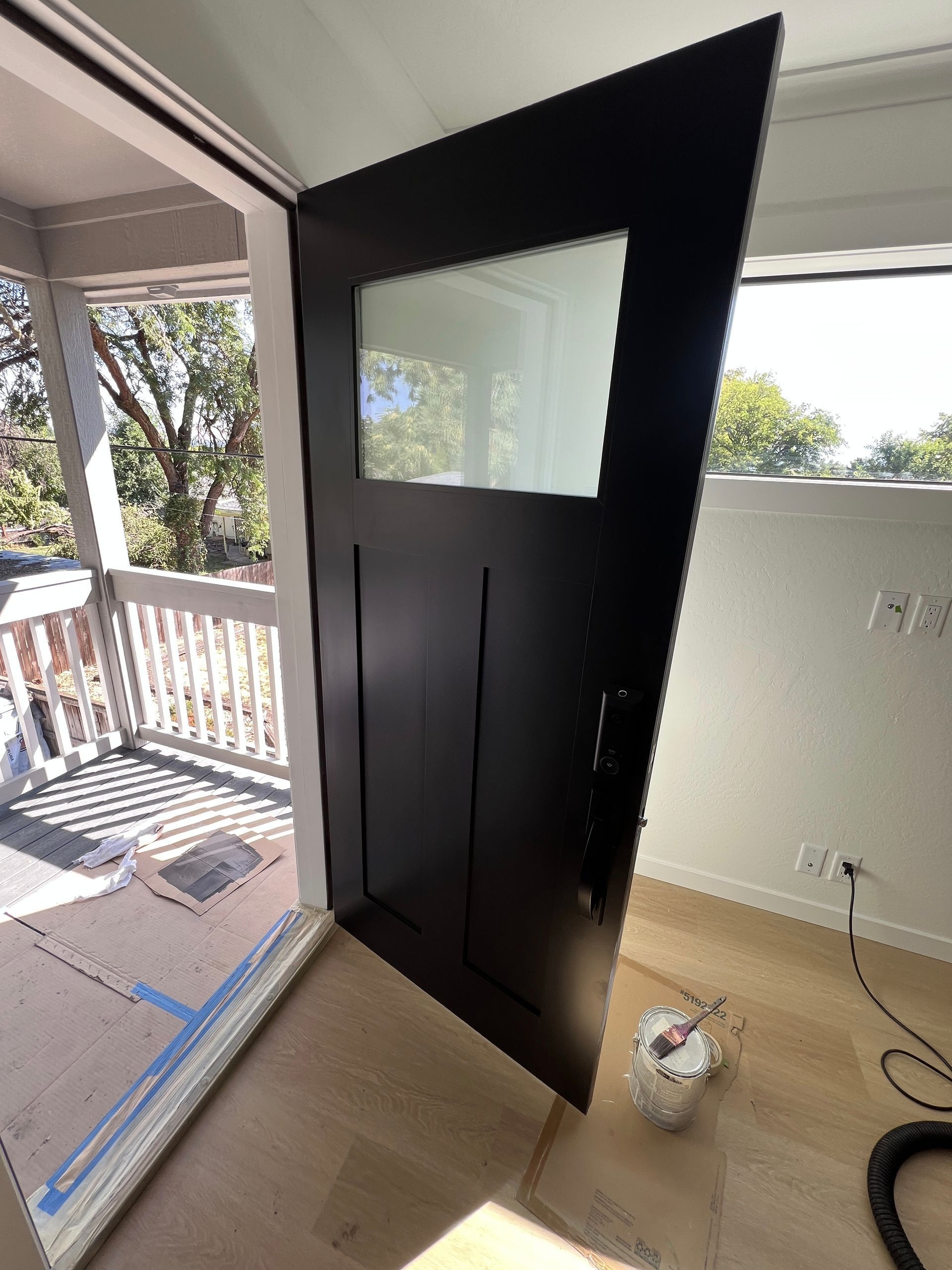 A modern dark brown door with a top glass panel stands open, revealing a sunlit wooden deck and an indoor room.
