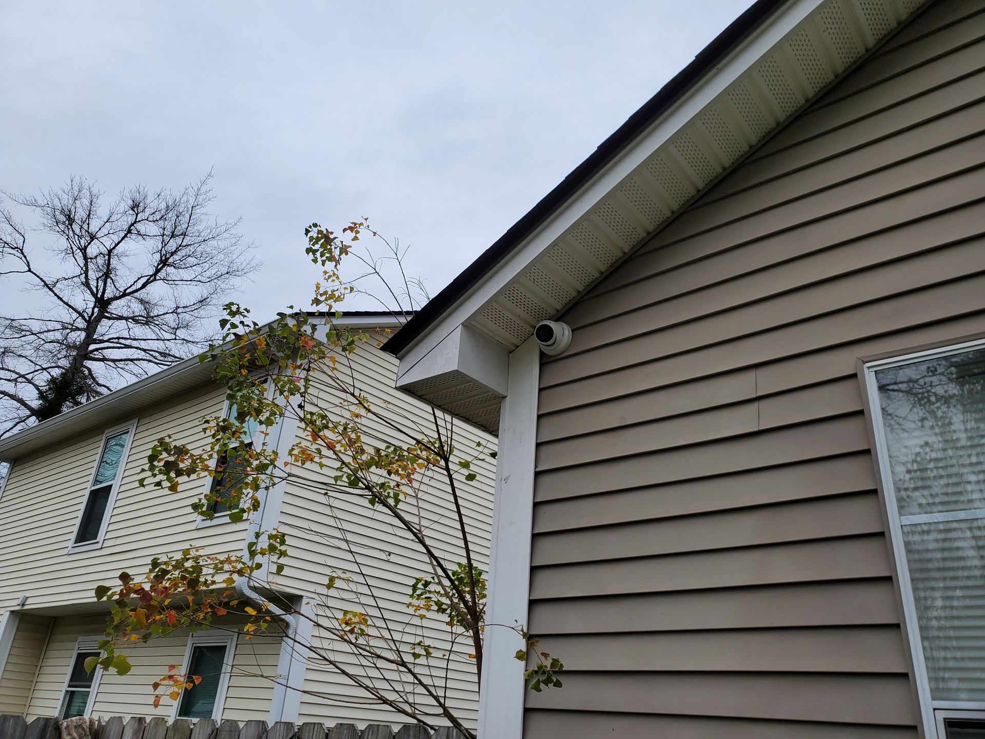 Two-story beige house exterior with white trim. A security camera is mounted under the eaves.