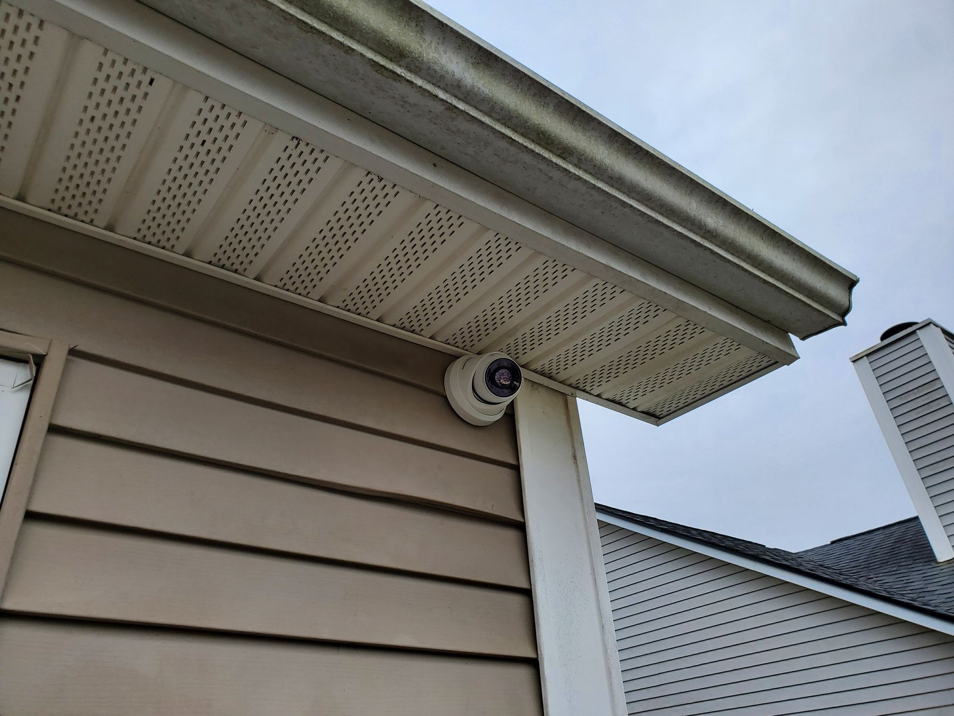 Security camera mounted under the eaves of a building with light brown siding and a white chimney in the background.