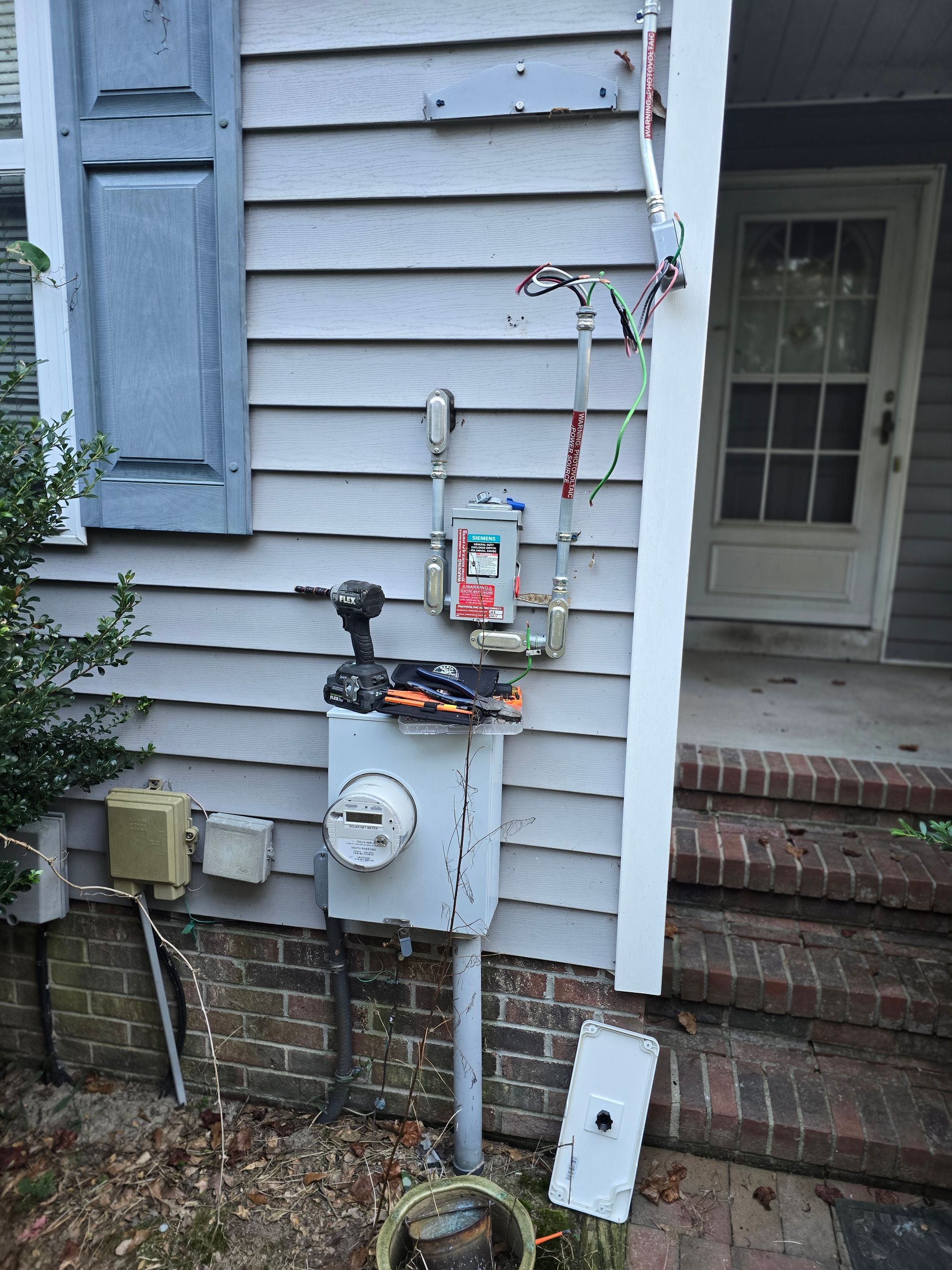 Exterior of house with electrical and gas meters, tools, and exposed wires. Gray siding, brick steps, and a white door.