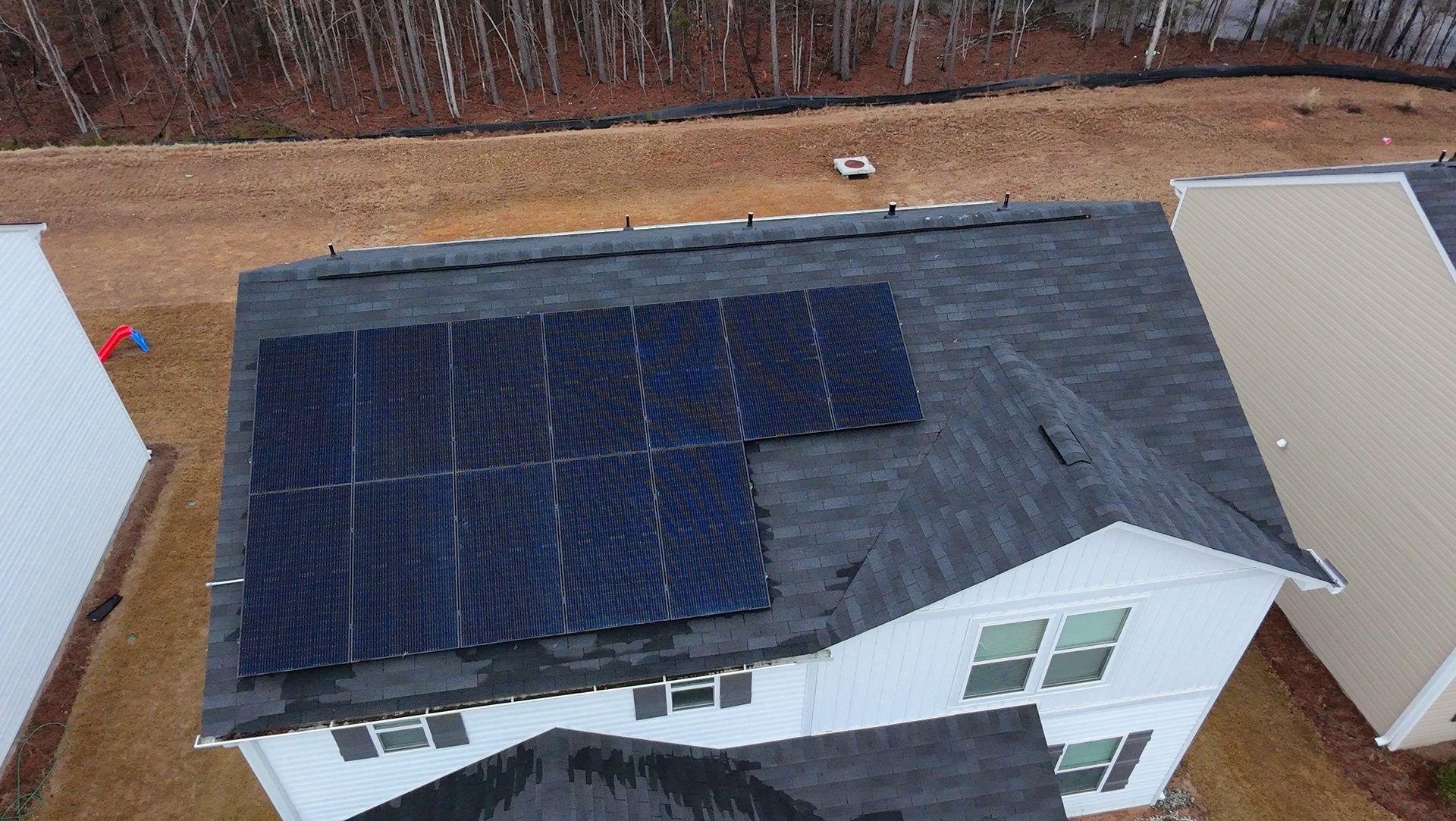 Solar panels on a gray shingled roof, trees cast shadows, with a paved driveway on the right.