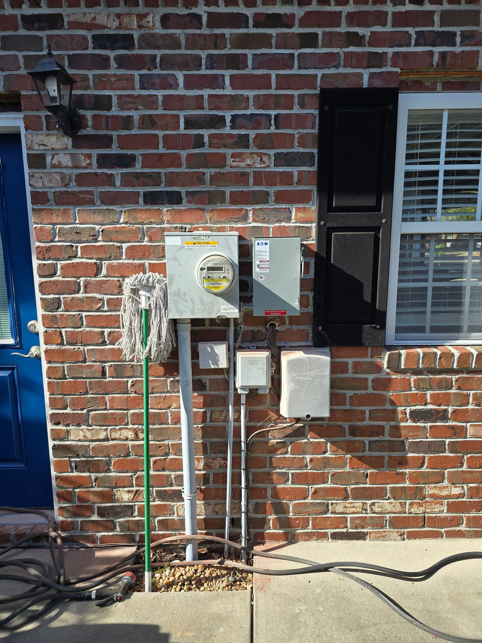 Brick wall with electrical meter and boxes, mop leaning against it, near a blue door and window.