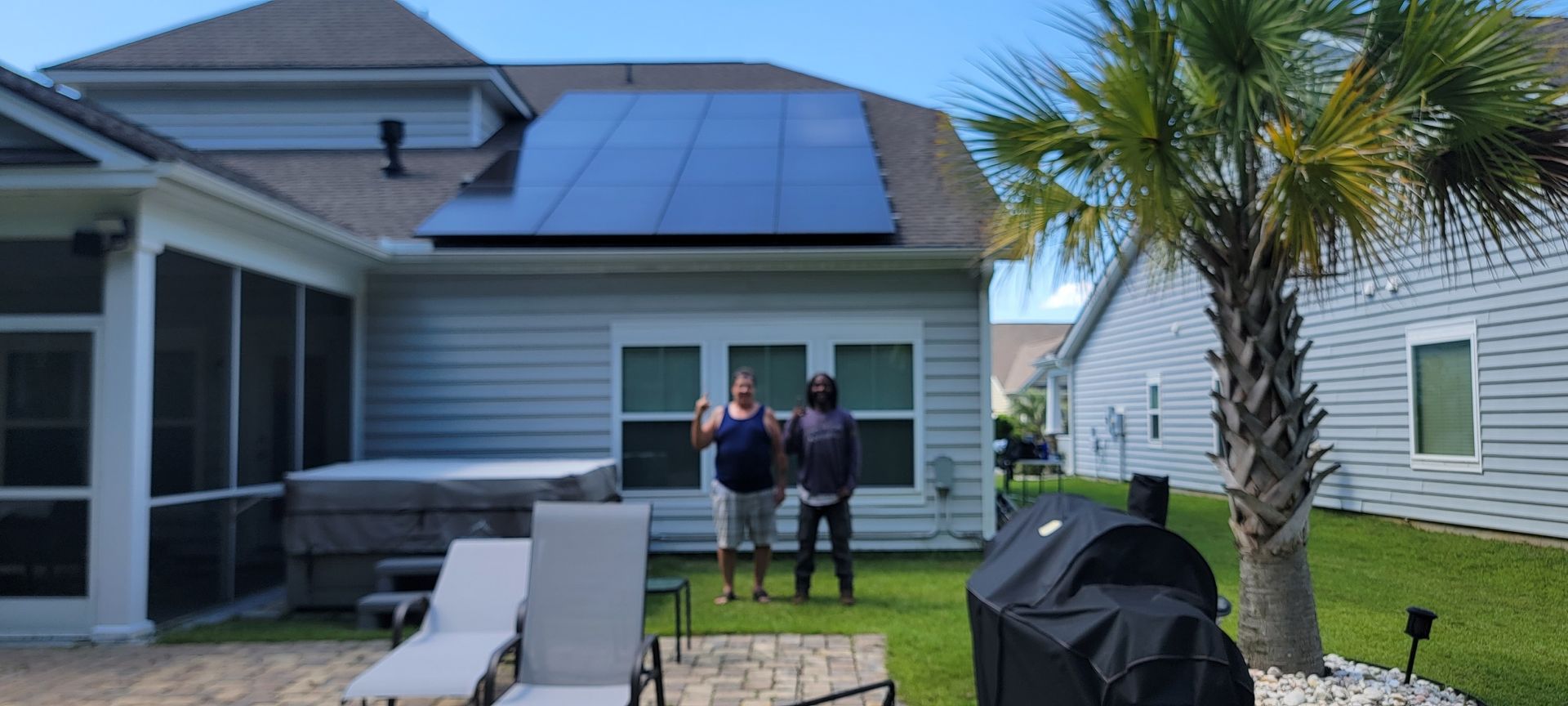 Two people stand in a backyard with a house, solar panels, and a palm tree.