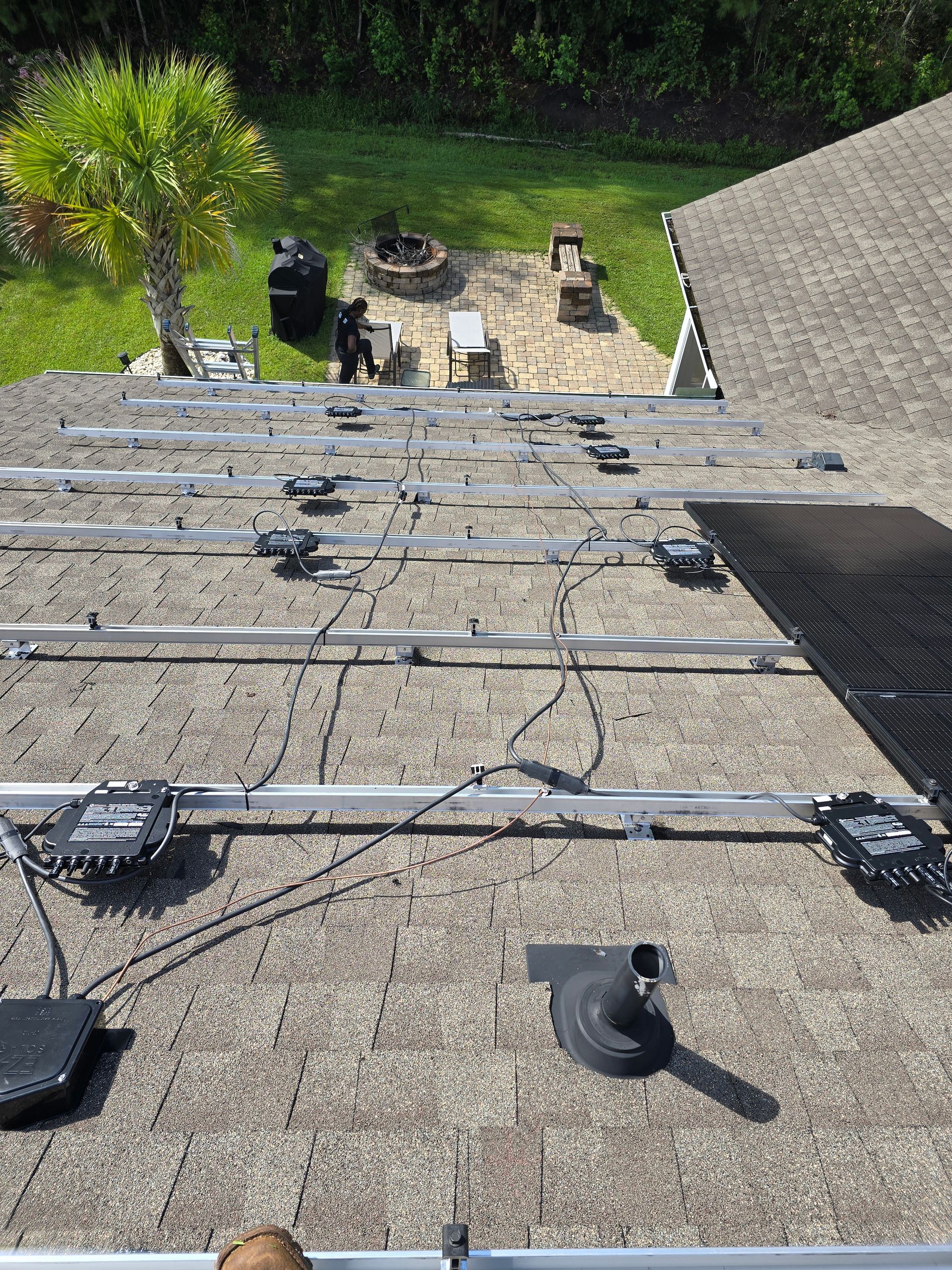 Solar panel installation on a rooftop with mounting rails and wiring. A person works near a backyard patio.