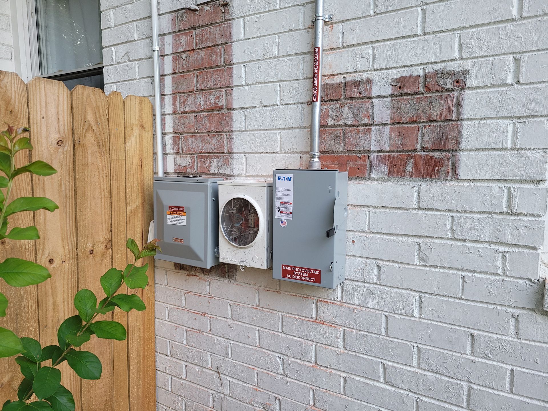 Electrical boxes mounted on a white brick wall next to a wooden fence and leafy green plant.