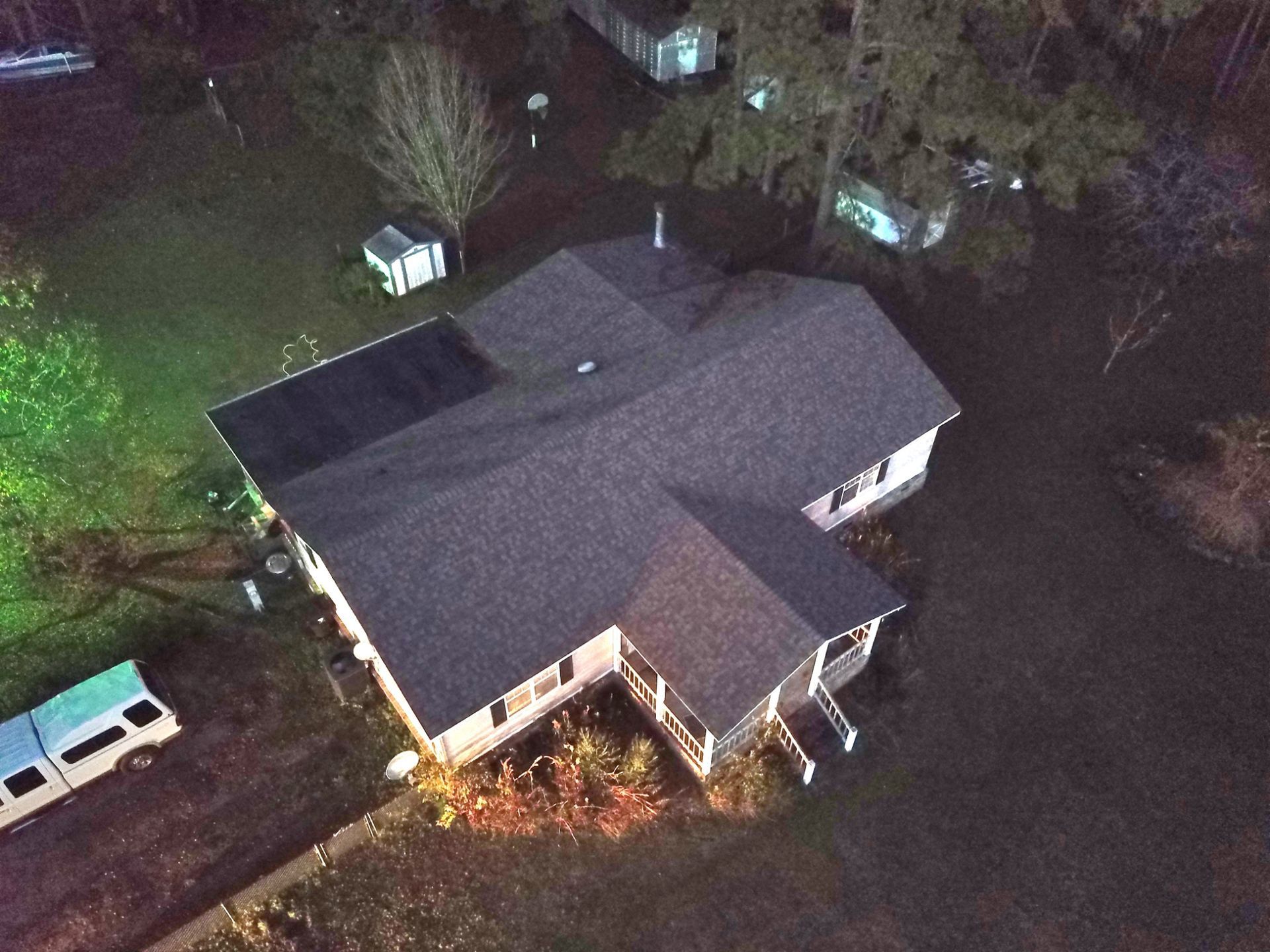 Overhead view of a house with a dark gray roof surrounded by trees and a lawn; a vehicle is parked nearby.