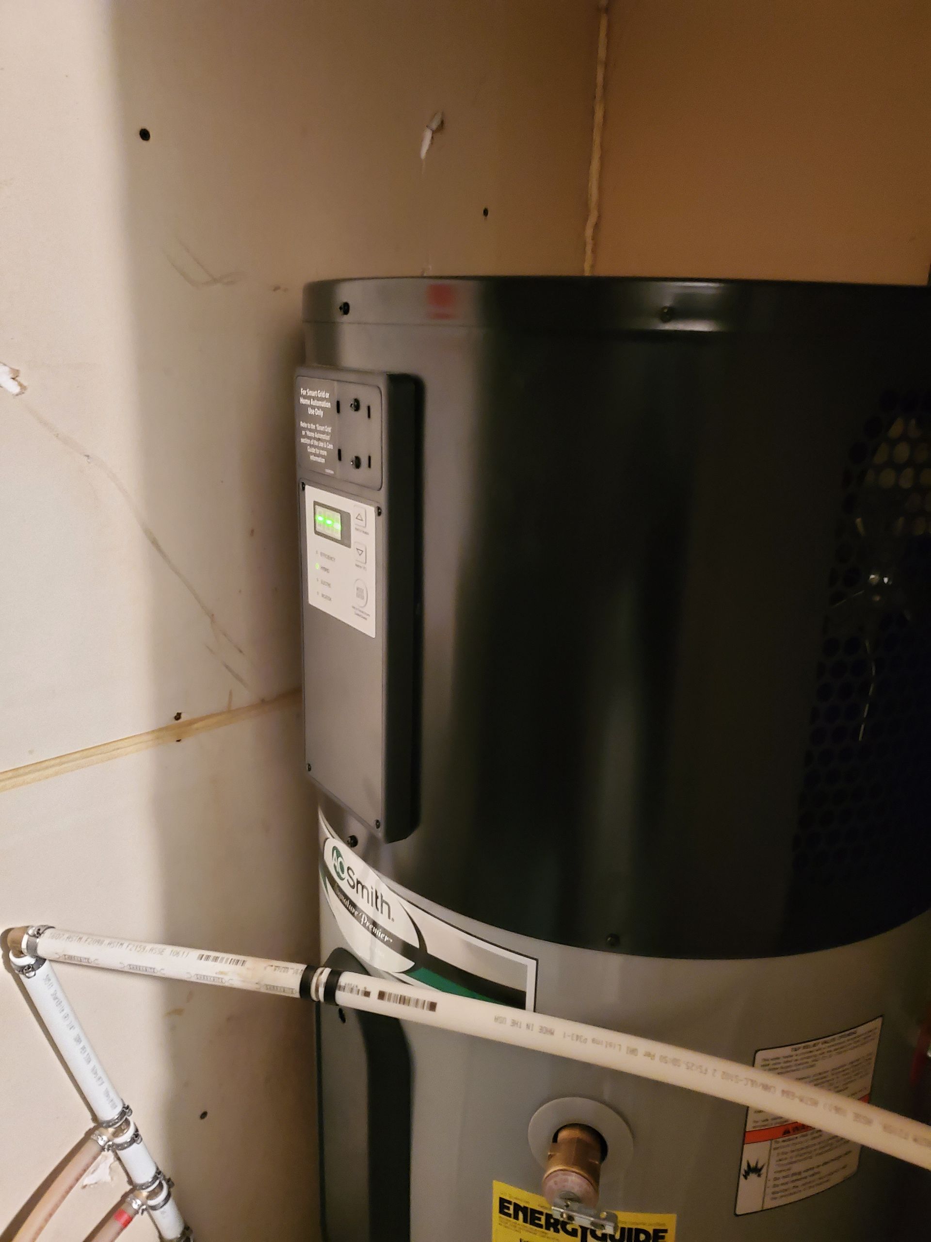 A water heater with a control panel is set inside a utility closet, with white pipes and walls nearby.