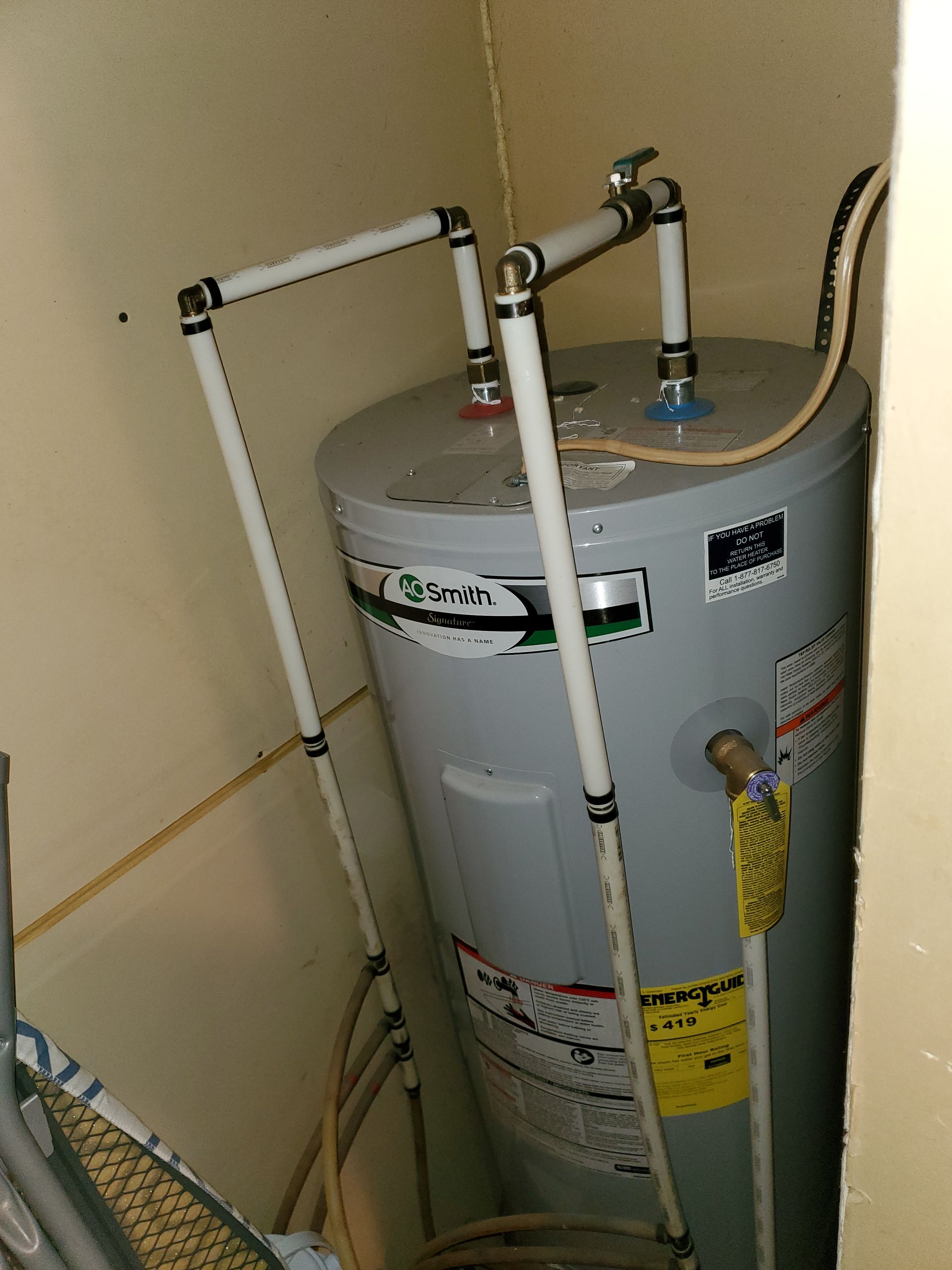 A water heater in a corner, with white pipes connecting. A yellow energy sticker is visible.