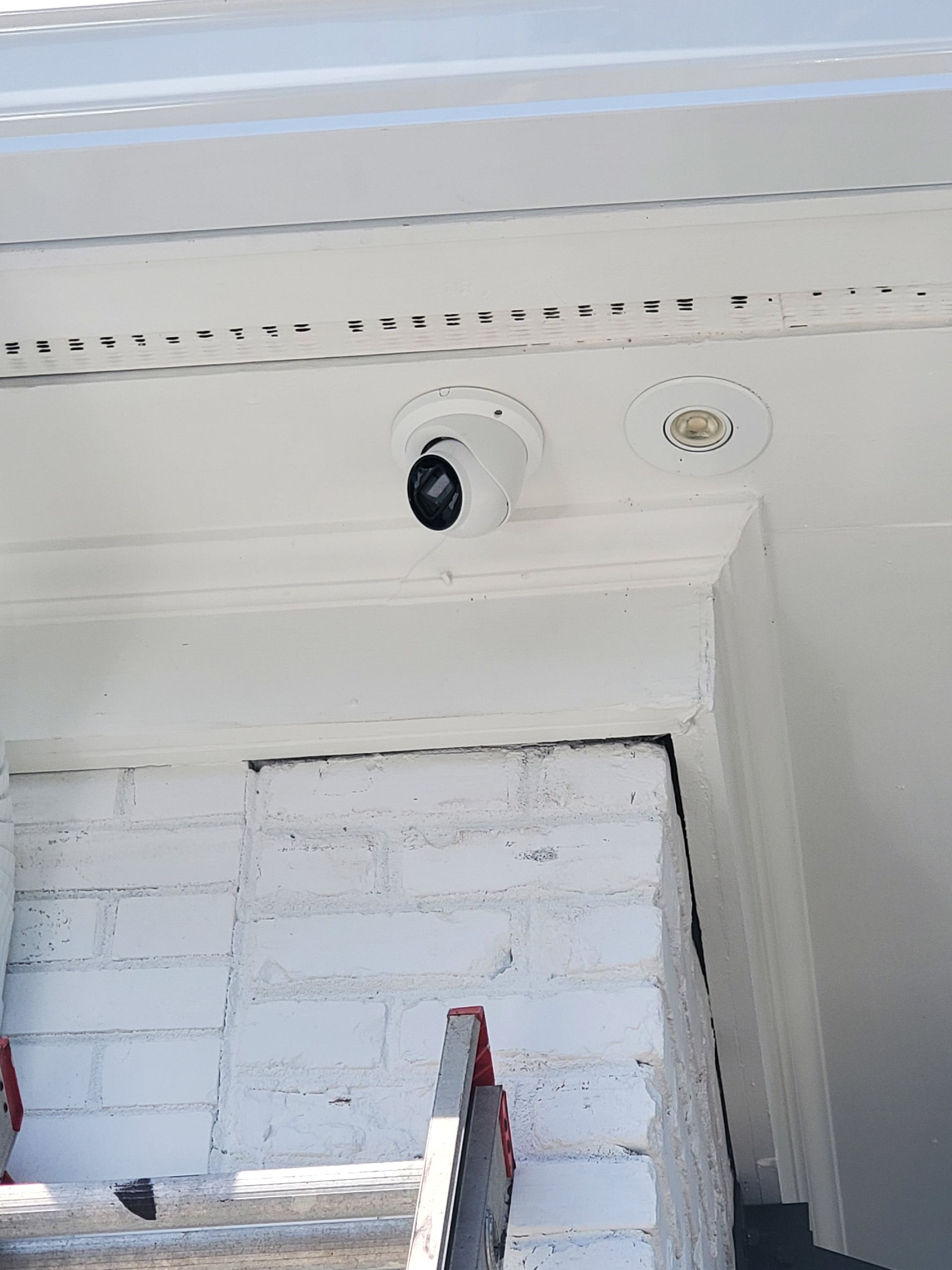 Security camera mounted on a white exterior ceiling, looking down at stairs and a brick wall.