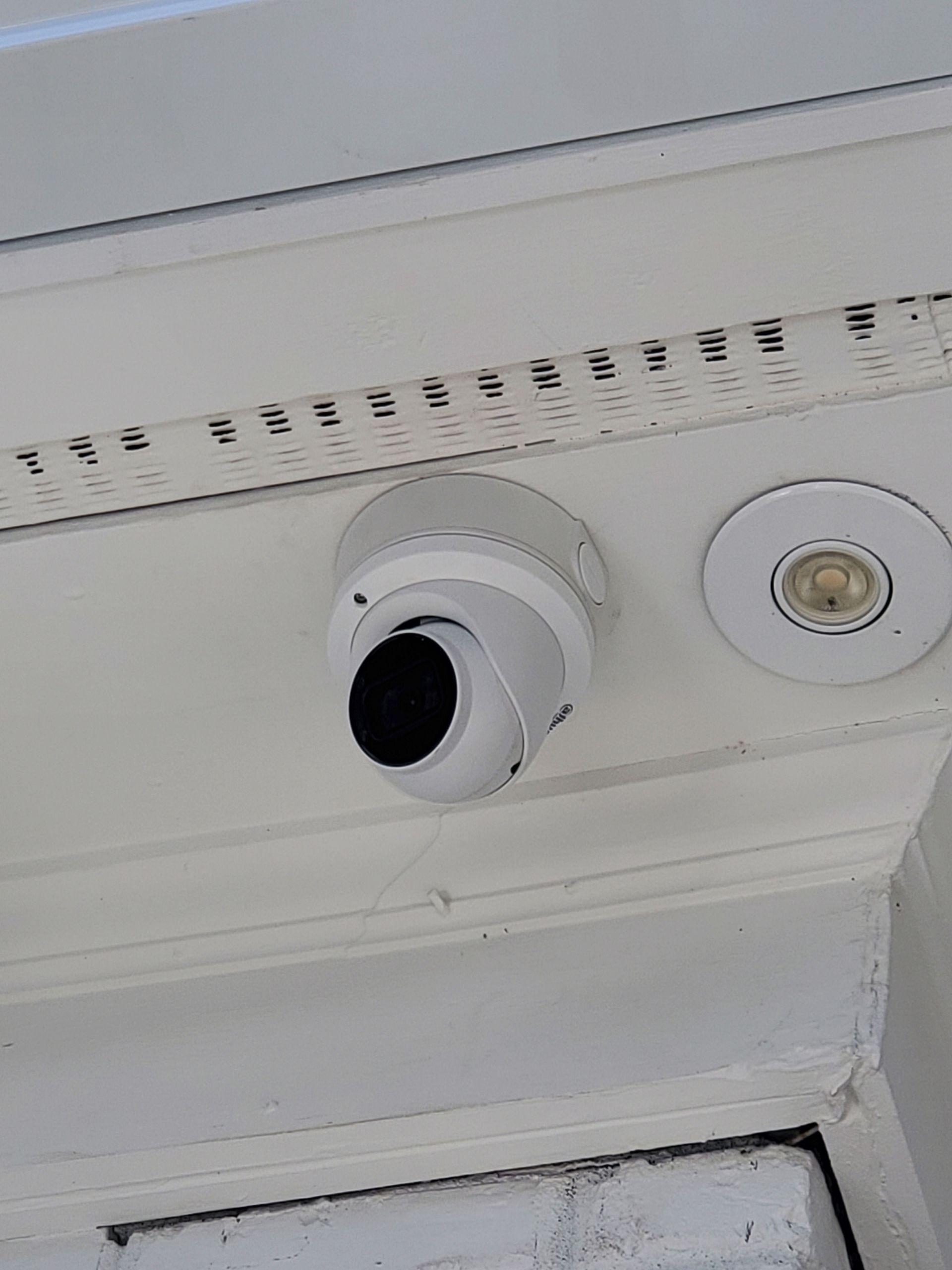 White dome security camera mounted on a white ceiling, next to a recessed light.