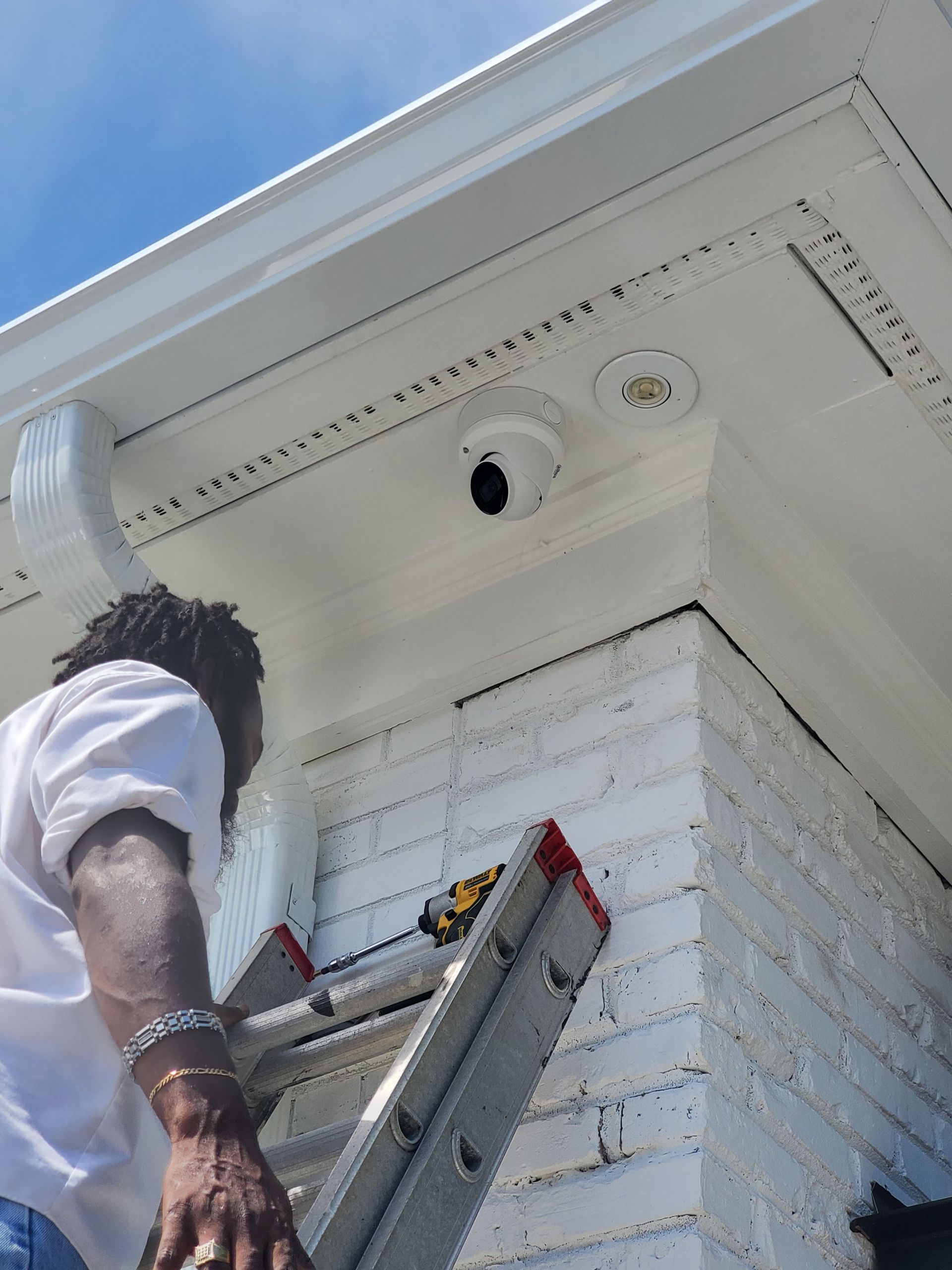 Person on a ladder installing a security camera on a white building's corner under a blue sky.
