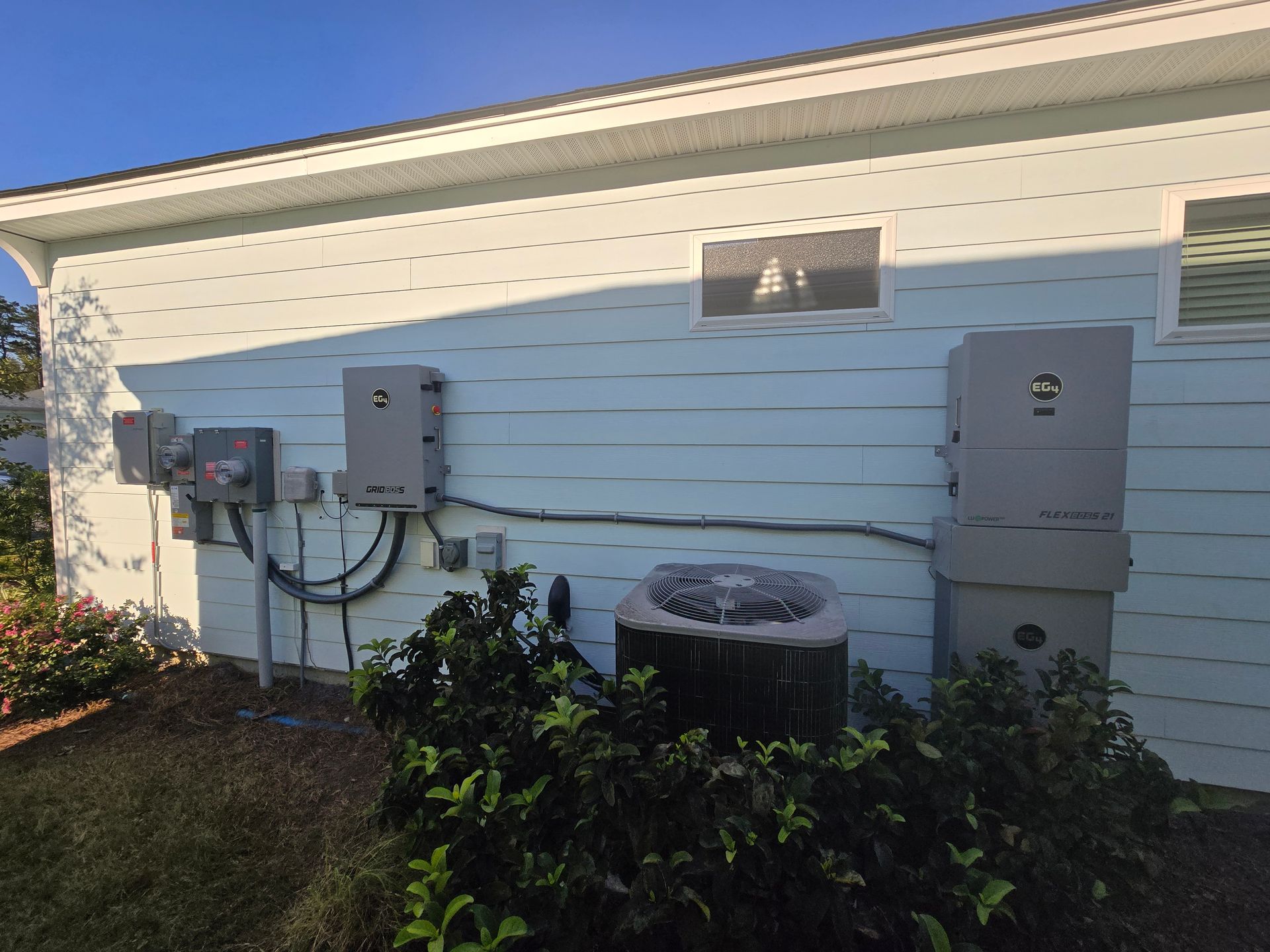 Solar panel inverters and electrical equipment mounted on the side of a light blue house, next to an AC unit.