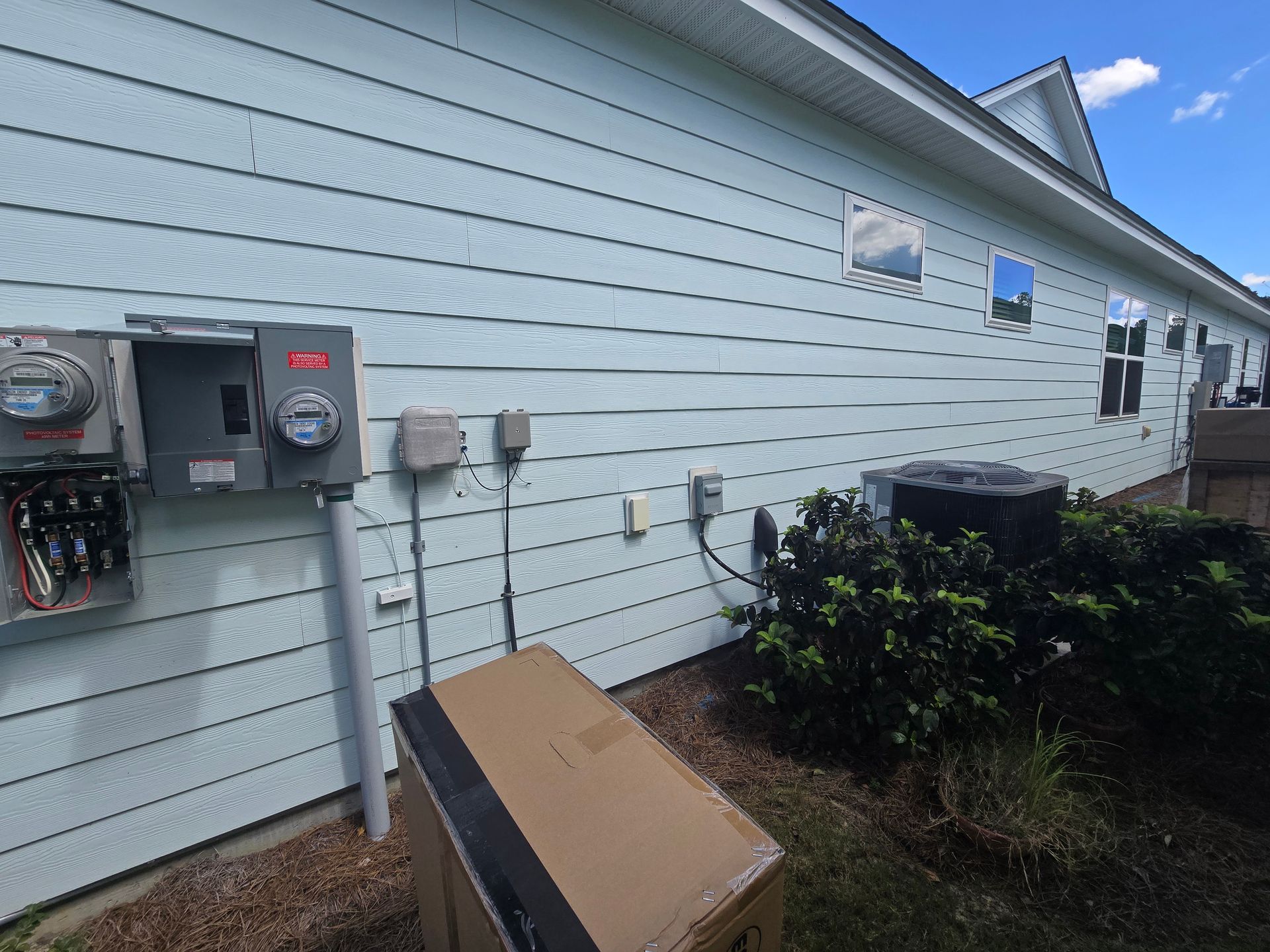 Exterior wall with electrical boxes, air conditioner, and a cardboard box on the grass.