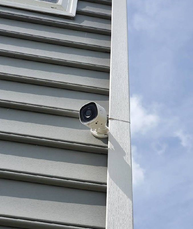 White security camera mounted on gray house siding, with a bright blue sky in the background.