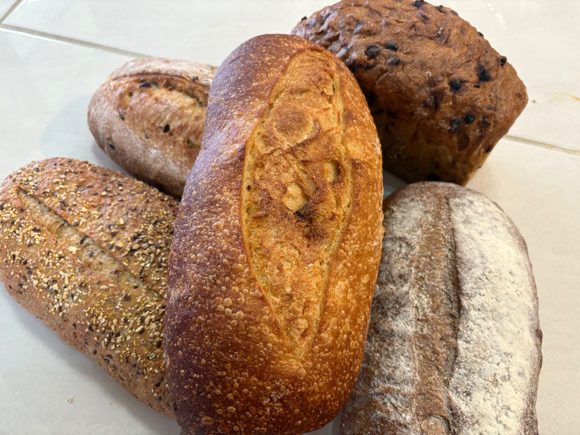 Assortment of five loaves of bread, with varied shapes, textures, and toppings, on a surface— Bread Basket in Kurri Kurri, NSW