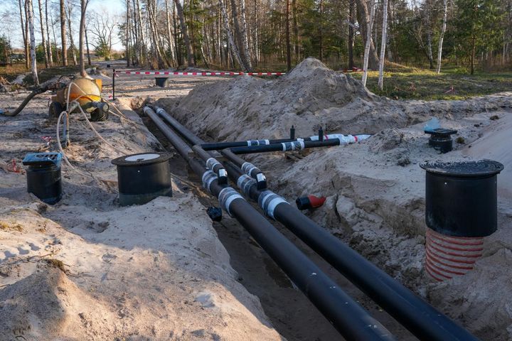Trenches with large black pipes and utility boxes at a construction site in a sandy wooded area