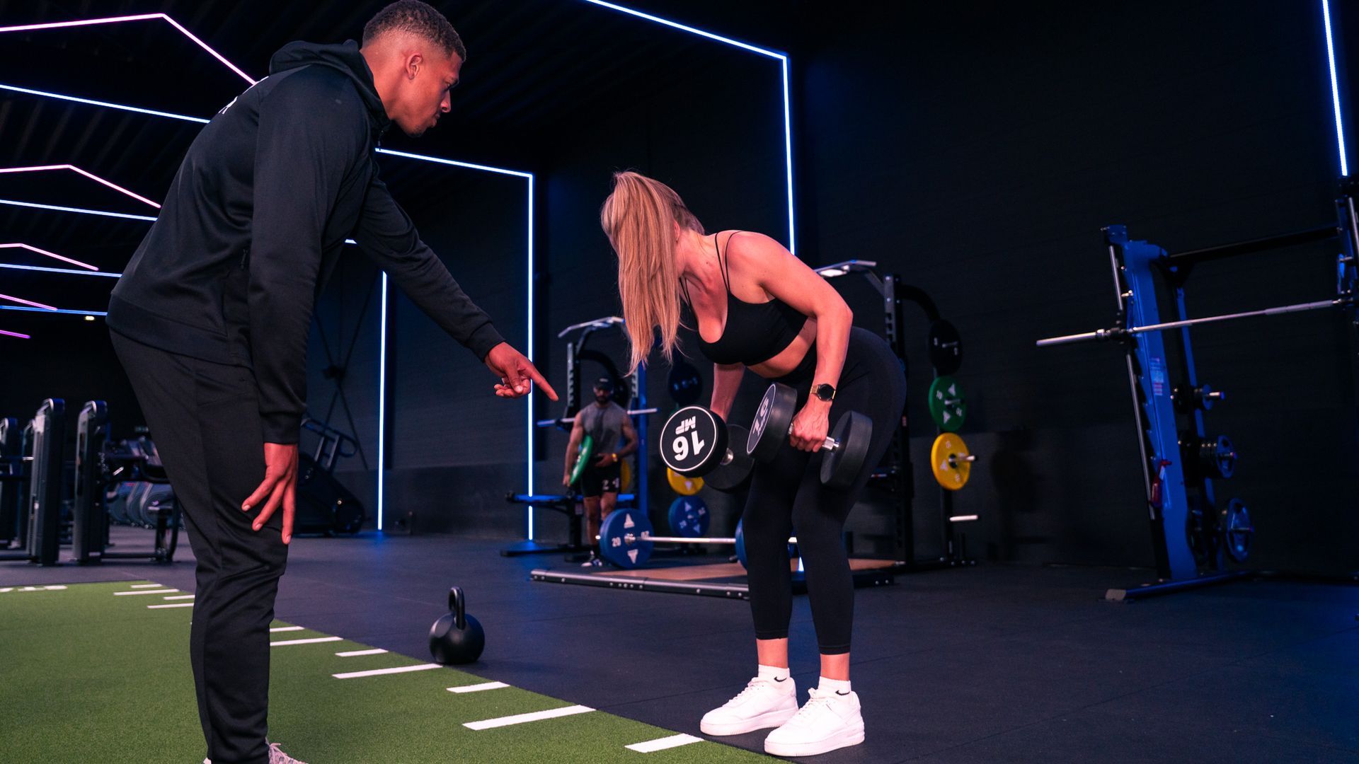 Een man wijst naar een vrouw die met dumbbells traint in een sportschool.