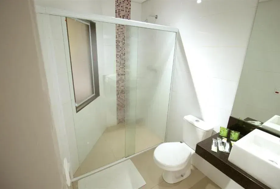 Banheiro com box de vidro, vaso sanitário, pia, espelho e uma pequena janela.