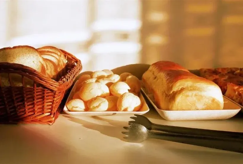 Pães em cestas e bandejas com pegadores de servir, dispostos em um balcão.