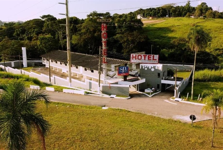 Prédio do hotel com placa, entrada e vagas de estacionamento, cercado por grama verde e árvores.