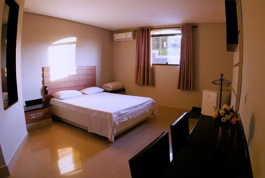 Quarto com cama, mesa de cabeceira, janela e escrivaninha escura. Uma luz quente ilumina o ambiente.