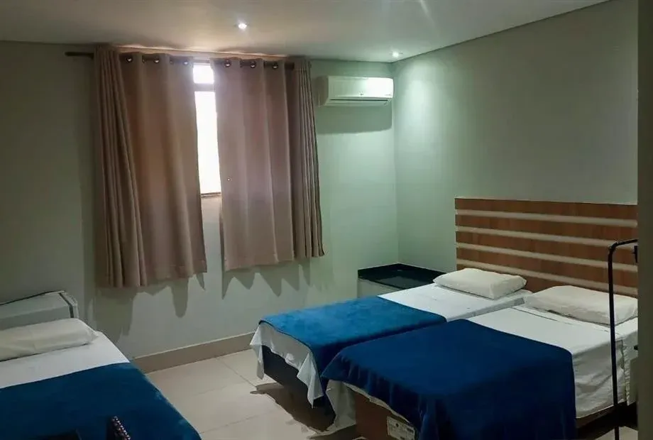 Quarto de hotel com três camas, janela com cortinas bege, paredes verde-claras e ar condicionado.
