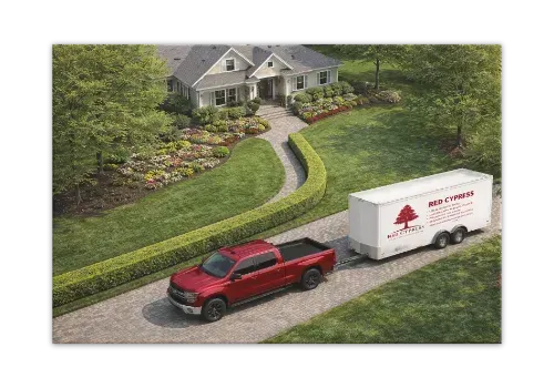 A red pickup truck is towing a white moving trailer up a gravel driveway toward a house surrounded by landscaping.