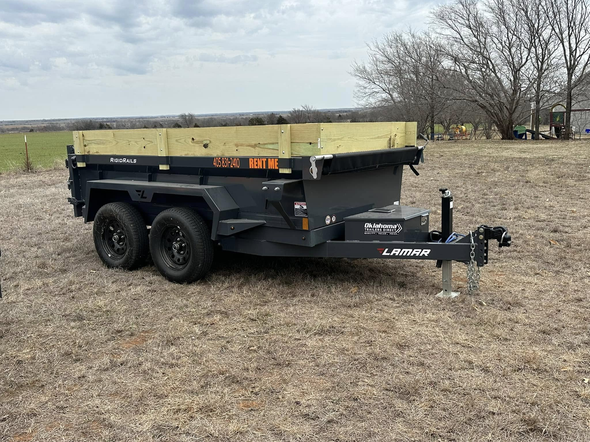 Black dump trailer with wooden sideboards on brown field, cloudy sky in background.