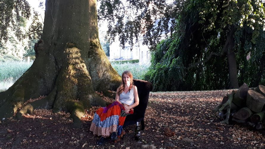 Vrouw zit in een zwarte stoel onder een grote boom met een kleurrijke rok; buitenomgeving.