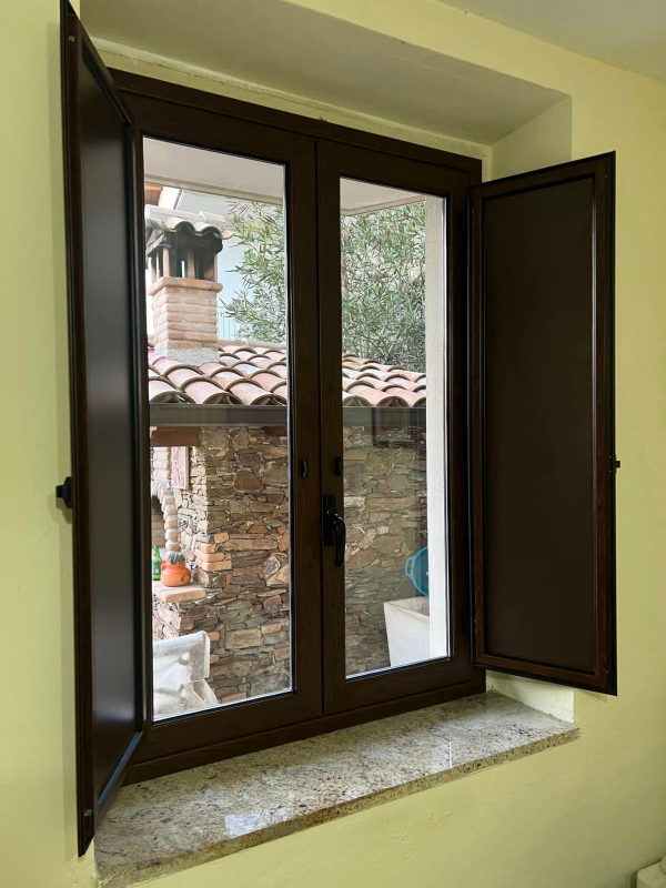 Finestra marrone con persiane aperte, che mostra la vista di una struttura in mattoni e tegole in terracotta.