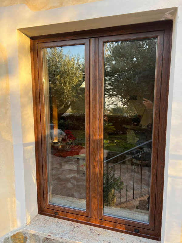 Doppia finestra con cornice marrone che riflette un giardino. Incastonata in una parete in stucco con davanzale in pietra.