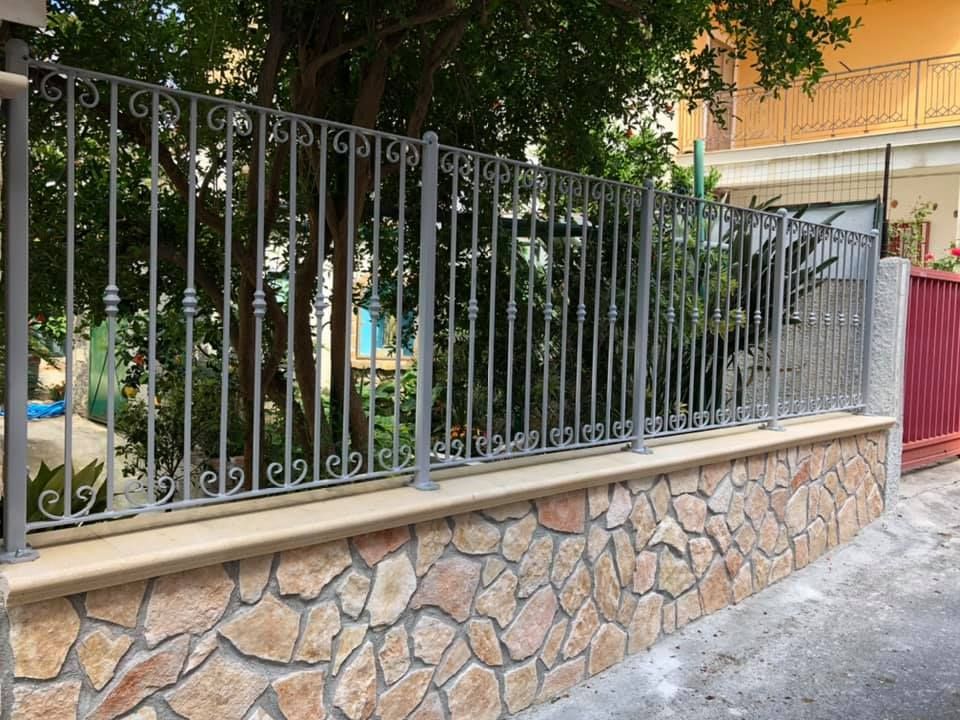 Muro di pietra con recinzione in ferro battuto di fronte a un albero e a un edificio.