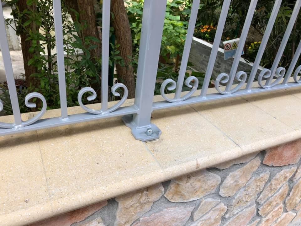 Ringhiera in metallo grigio con volute decorative su un muro in pietra beige con fondamenta in roccia, all'aperto.