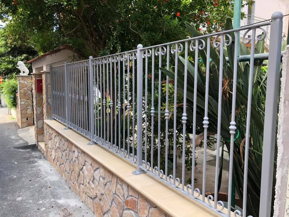 Recinzione in metallo grigio sopra un muro di pietra lungo un marciapiede con alberi sullo sfondo.