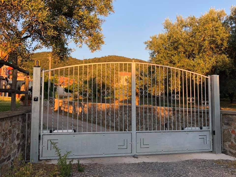 Cancello carrabile in metallo argentato, parte superiore ad arco, sbarre verticali, con apertura elettronica, incastonato in un muro di pietra.