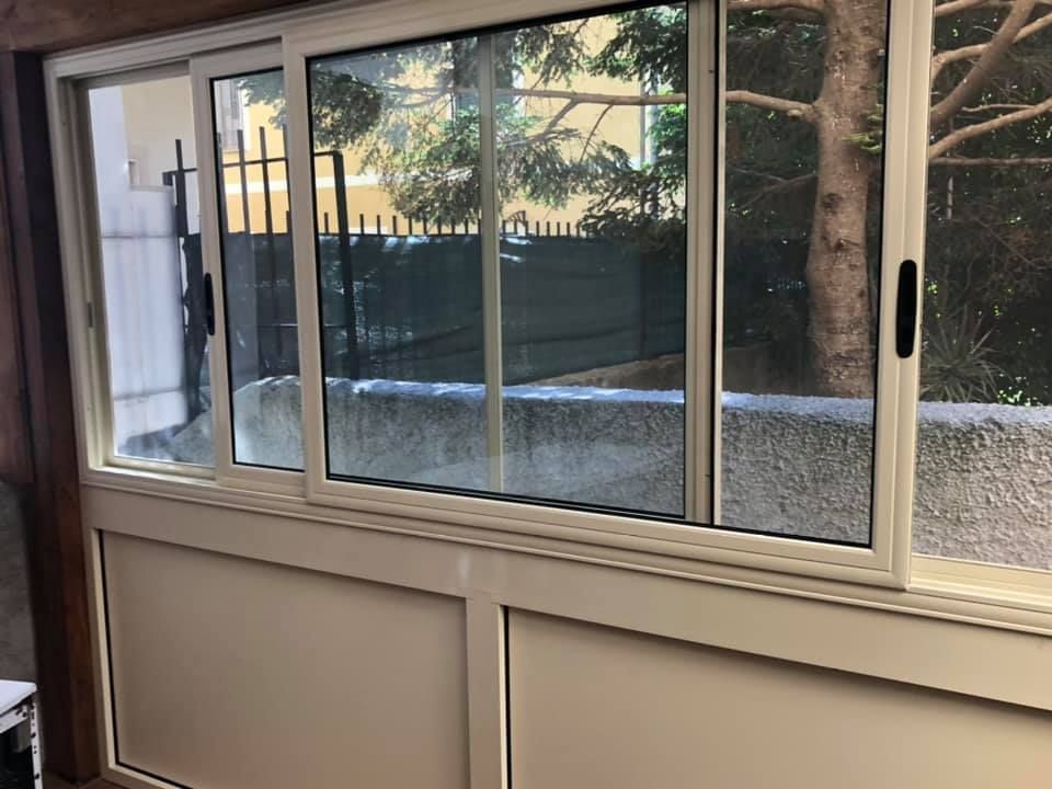 Finestre scorrevoli in vetro con cornici bianche che si affacciano su un cortile con alberi e un muretto basso.