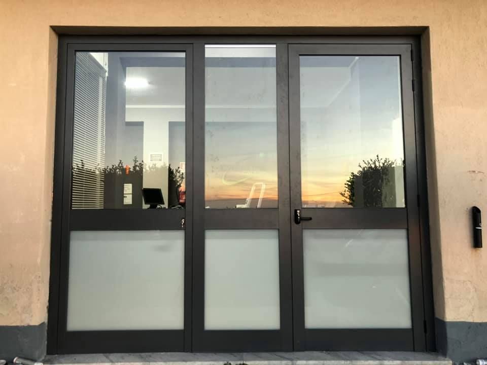 Porta in vetro a tre ante con pannelli inferiori smerigliati. Telaio grigio scuro. Vista esterna con riflesso del tramonto.