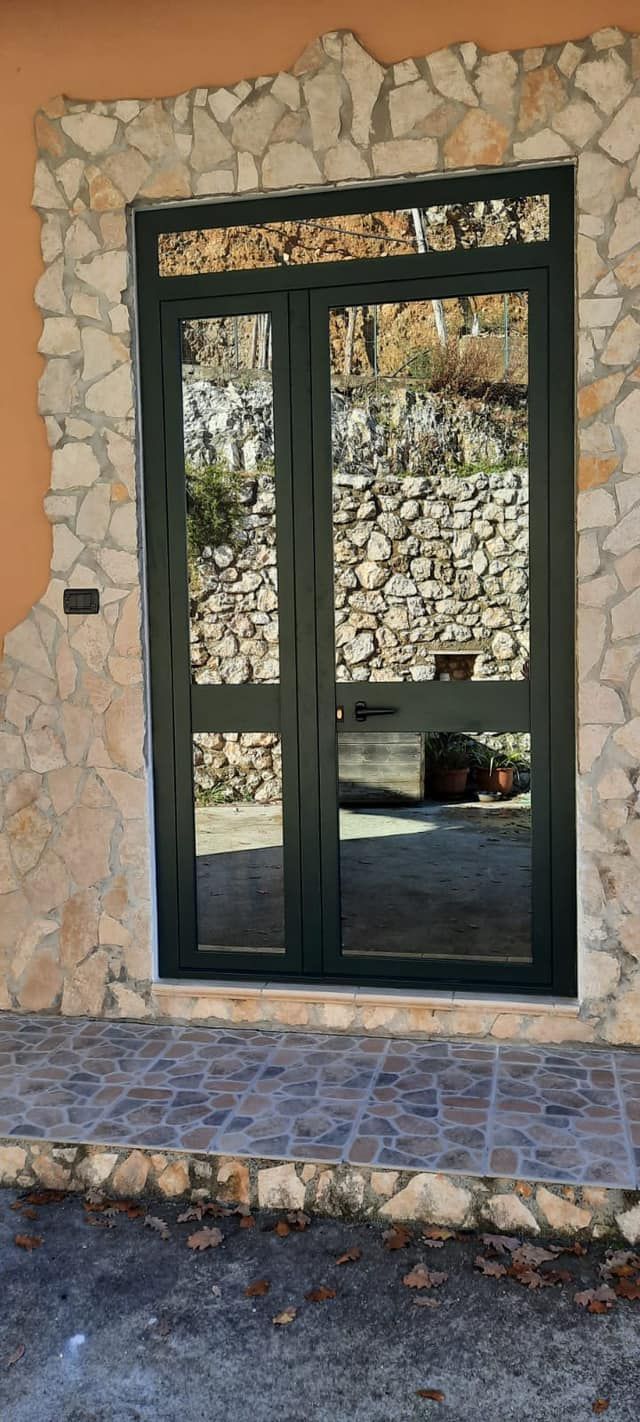 Porte doppie verde scuro con pannelli in vetro, circondate da un muro in pietra e intonaco arancione. Patio piastrellato grigio.