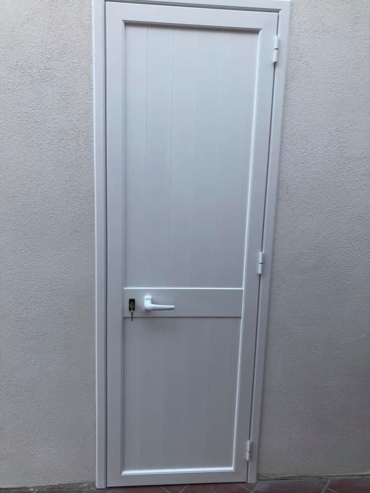 Porta bianca rettangolare con maniglia e serratura contro una parete chiara e ruvida.