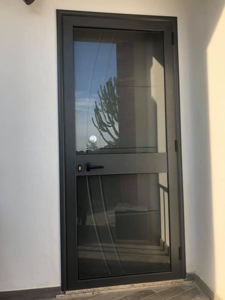 Porta grigio scuro con zanzariera, incorniciata da una parete bianca. Riflesso del paesaggio esterno sul vetro.