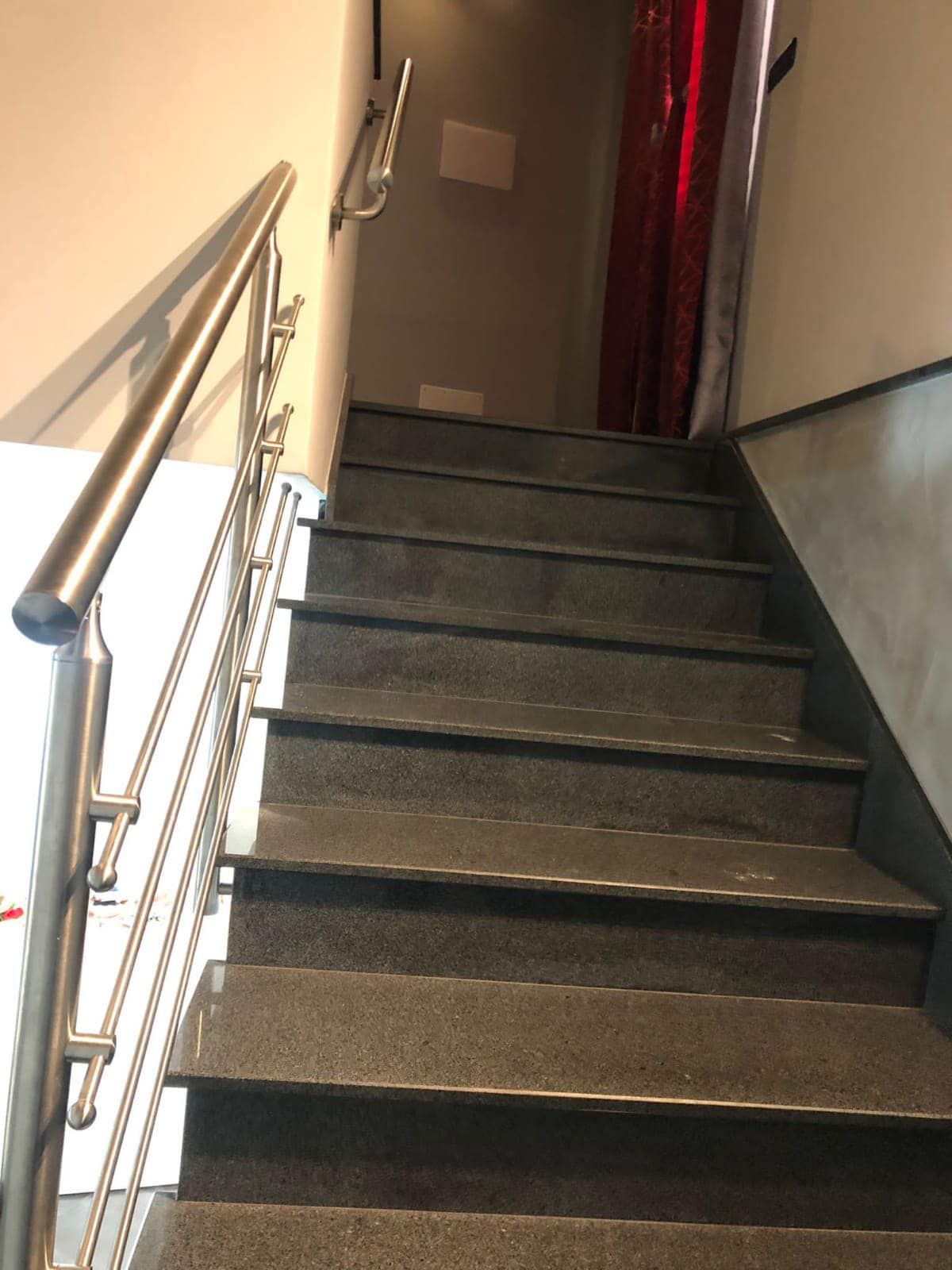 Scala con corrimano in acciaio inox che sale. Gradini grigi, pareti chiare e una tenda rossa visibile.