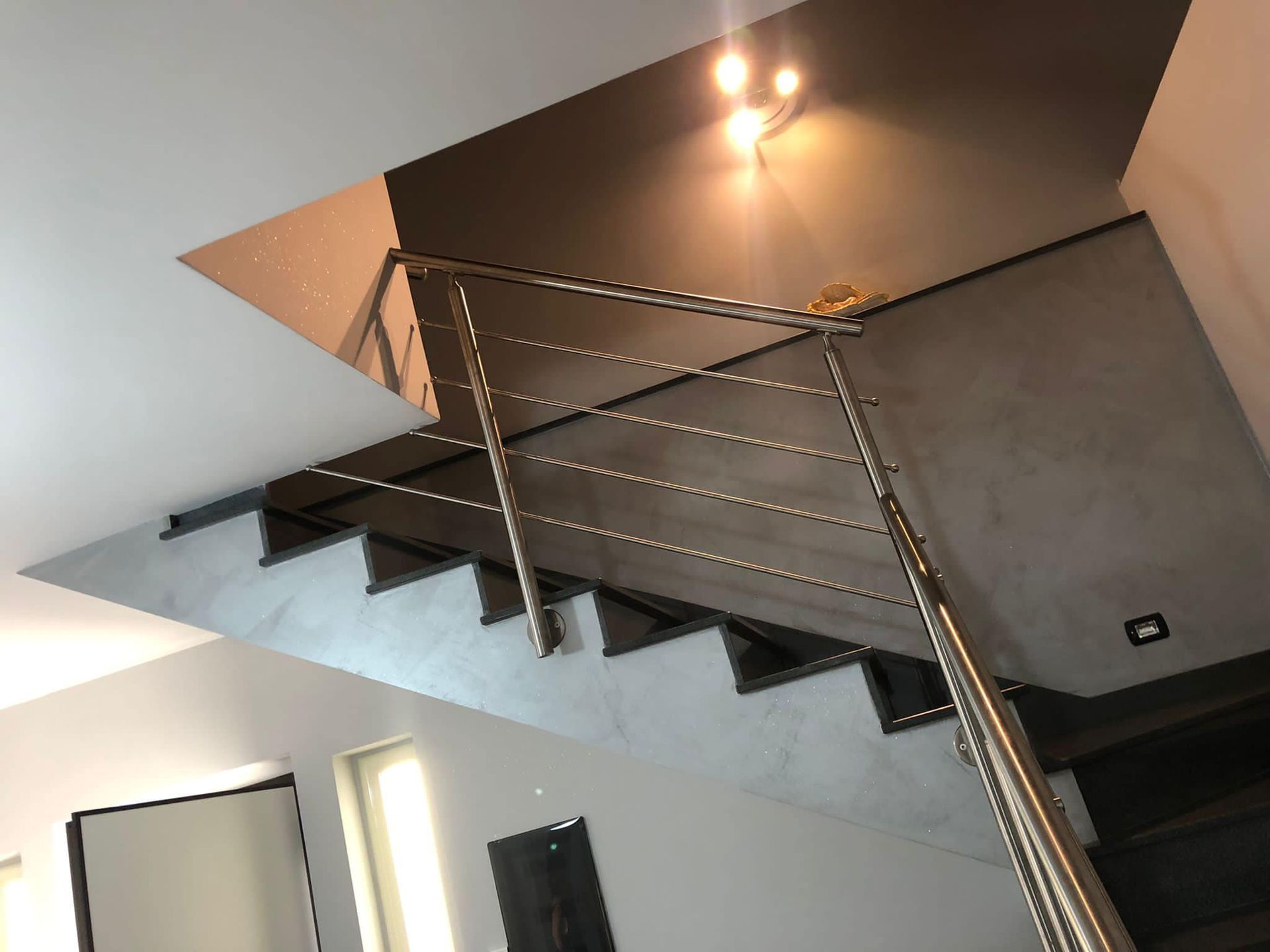 Scala con ringhiera in acciaio inox e gradini scuri su pareti grigie e bianche.